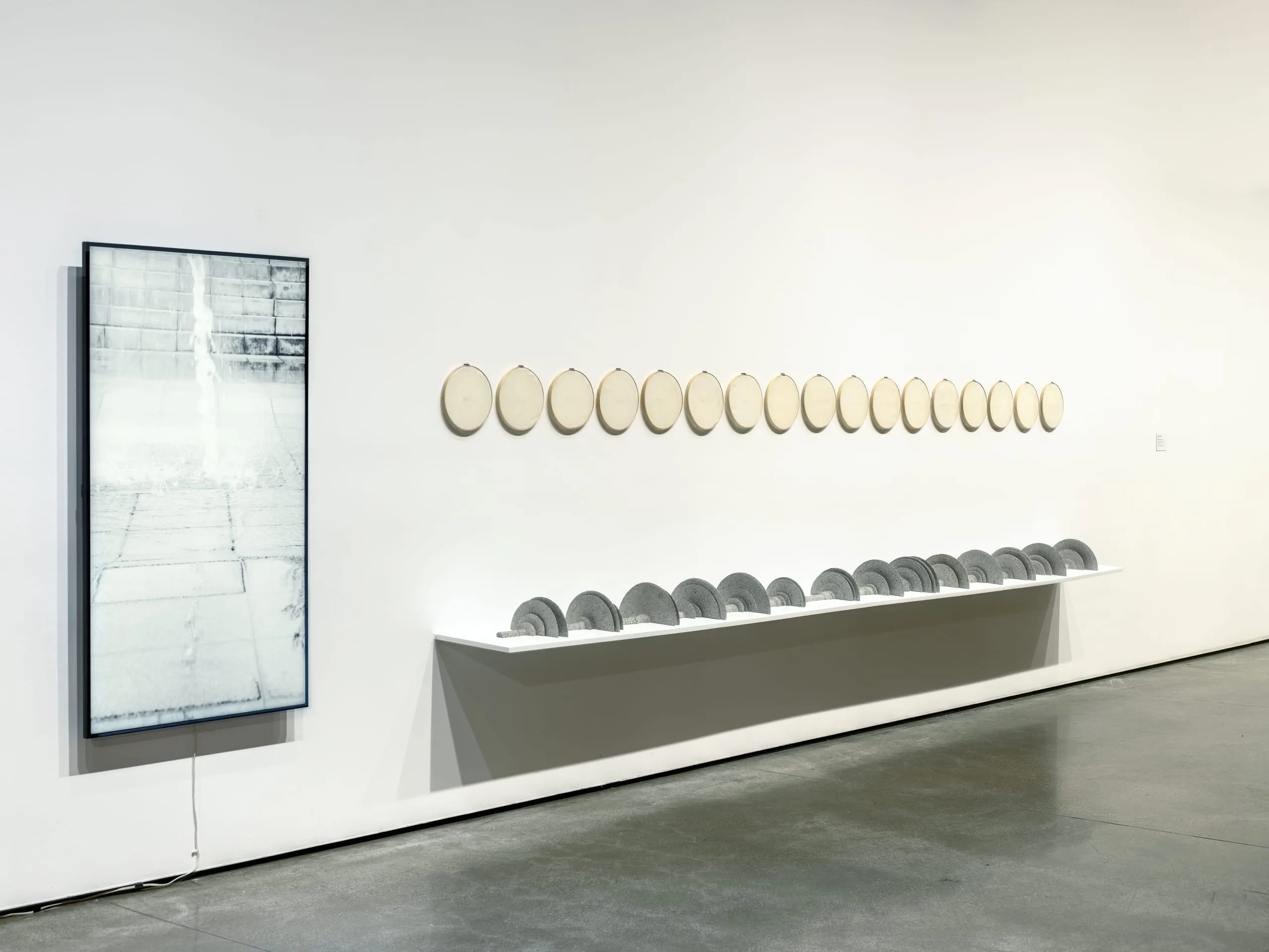 Gallery installation with two components: a vertically oriented light box photograph in a dark frame showing a pale, abstracted architectural scene, and a long white shelf holding a row of gray ceramic disc forms, above which a series of oval textile-covered hoops hang in a horizontal line along the white wall.